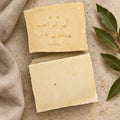 Two Laurel Aleppo soap bars on warm limestone with beige linen and a laurel sprig
